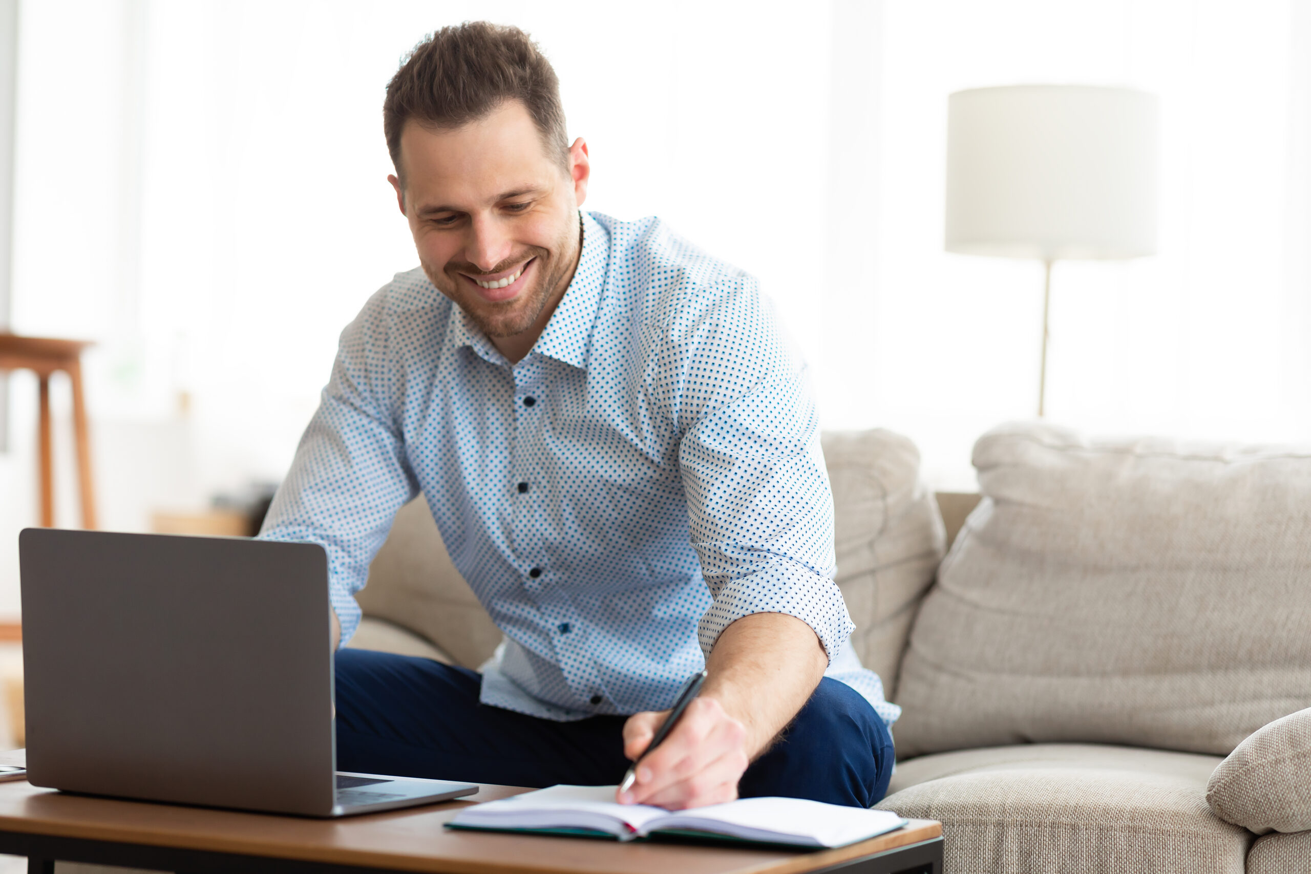 Tips For Writing The Best Social Media Copy Note Taking Concept. Smiling handsome man using laptop, writing in notebook, sitting on couch, copy space